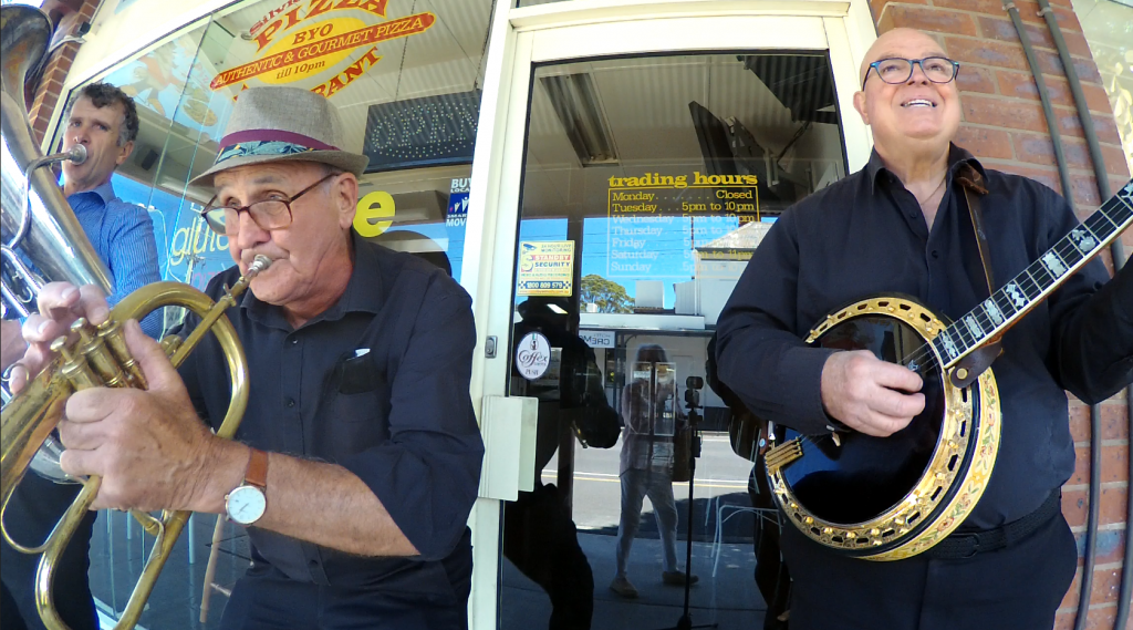 A tuba, flugel horn and banjo players have fun in front of a pizza shop.